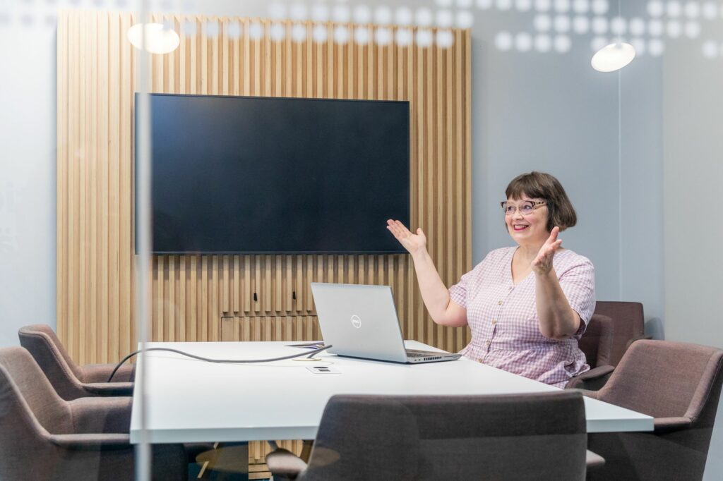 Sanna Vehviläinen ojentaa käsiään ylöspäin ja istuu neuvotteluhuoneessa läppäri edessä.