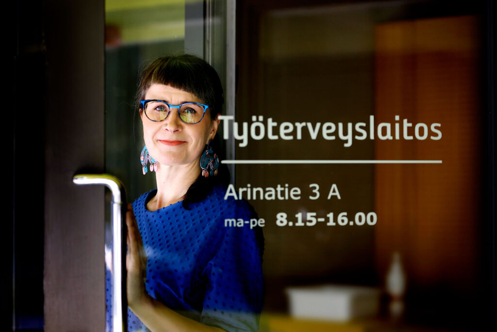Heli Clottes Heikkilä seisoo Työterveyslaitoksen oven takana ja katsoo kameraan.