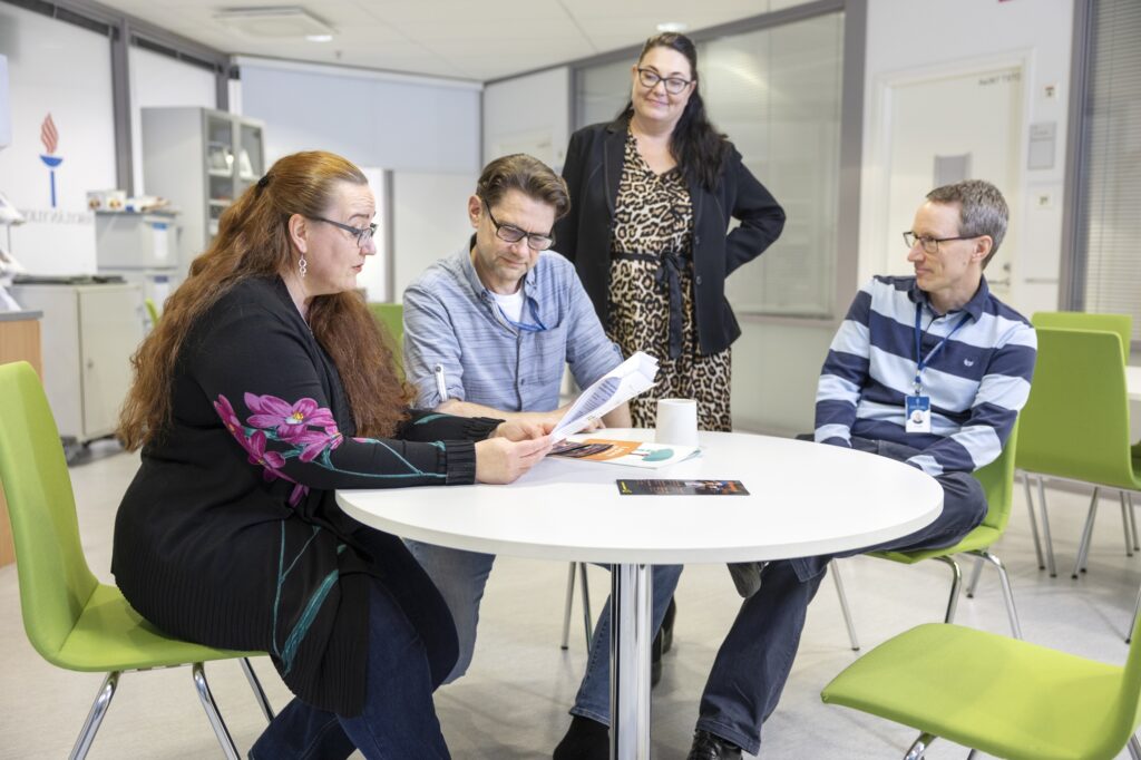 Johanna Rantanen ja kolme työkaveria taukotilassa Jyväskylän yliopistossa. Johanna ja kaksi työkaveria istuvat pöydän ääressä ja katsovat monisteita ja yksi työkaveri seisoo taustalla.