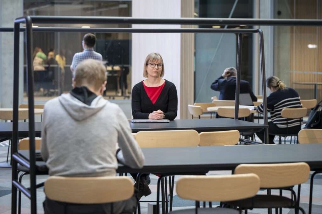 Jenni Radun istuu pöydän takana ja katsoo kameraan. Taustalla näkyy opiskelijoita. Jenni Radun kuvattu Turun AMK:n EduCityn tiloissa avoimissa oppimistiloissa.