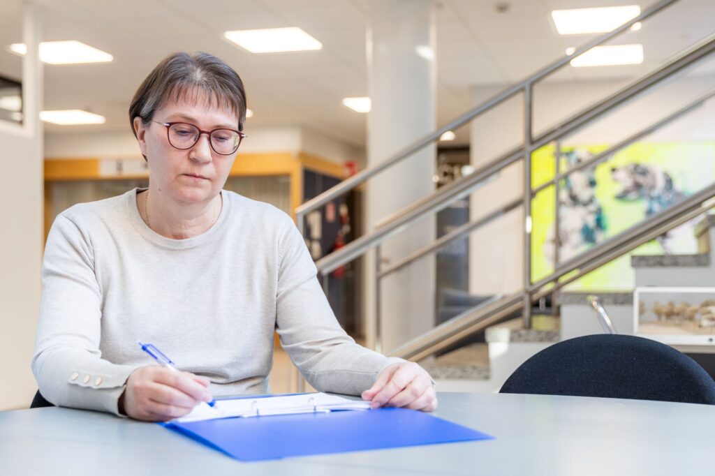Sirpa Rautiala istuu pöydän ääressä ja katsoo alaspäin pöydällä olevaa työpaperia. Oikeassa kädessä hänellä on kynä. Taustalla näkyy portaat ja kaide sekä eteistilaa.