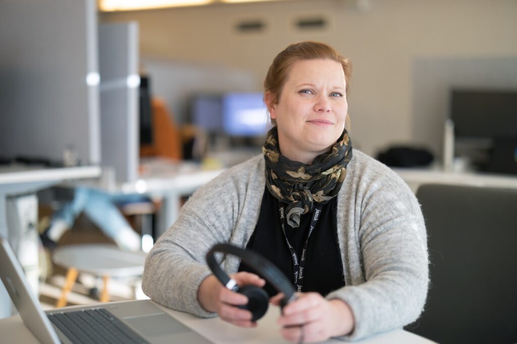 Tutkija Anna-Liisa Ojala kuvattuna Jyväskylässä JAMK:n pääkampuksella Rajakadulla. Anna-Liisa istuu ja pöydällä on läppäri ja kädessä kuulokksseet. Taustalla näkyy tietokoneita.