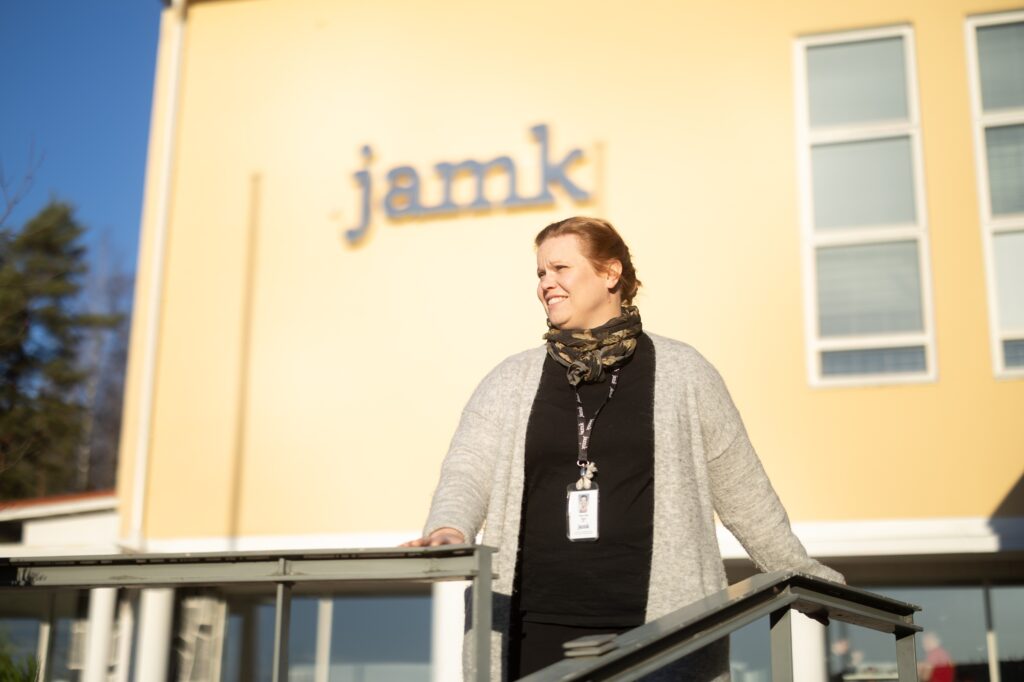 Tutkija Anna-Liisa Ojala kuvattuna Jyväskylässä JAMK:n pääkampuksen ulkopuolella Rajakadulla. Taustalla näkyy pääkampuksen rakennus, jossa lukee jamk. Anna-Liisa nojaa kaiteeseen ja ktasoo sivuviistoon.