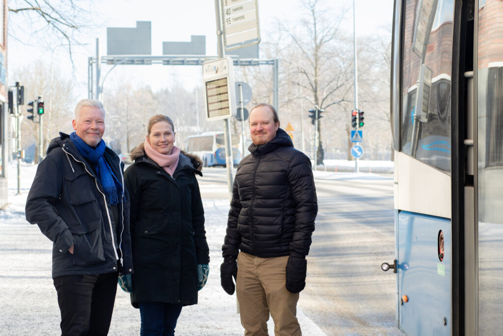 Henkilökuvassa professori Petri Nokelainen sekä tutkijat Eija Lehtonen ja Ilmari Puhakka. Oikealla näkyy bussin takaosa ja taustalla liikennevaloja ja tie.
