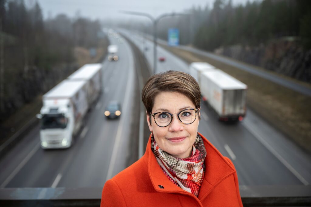 Susanna Visuri kuvassa etualalla ja taustalla näkyy maantietä, jossa ajaa rekka-autoja ja henkilöautoja. Taustalla sivulla näkyy piennarta ja metsää.
