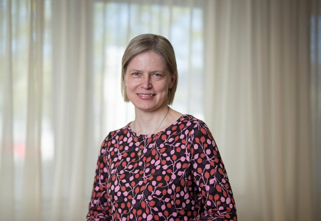 Professori Minna Stolt lähikuvassa. Takana näkyy vaaleat verhot.